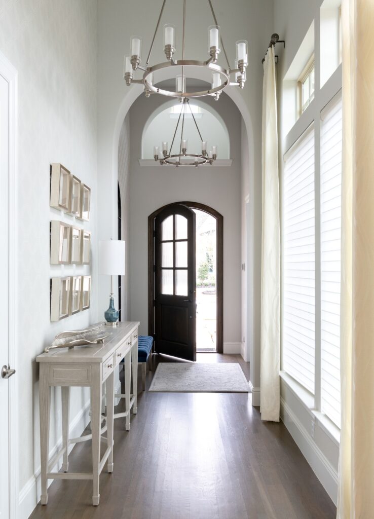 An Arched Mirror Above A Navy Plush Bench Reflects Light Beautifully, Creating An Airy Welcome. The Arches Continue Through The Foyer As You Enter The Front Door. The Circular Silhouette Of The Chandeliers Keeps The Lines Soft While Drawing You Into The Home. Touches Of Blue From The Bench To The Console Lighting Offer A Refreshing Yet Calming Pop Against The Neutral Palette.